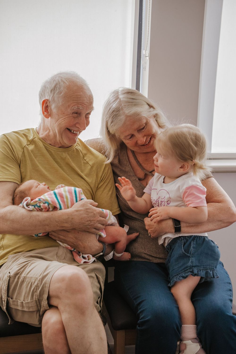 Grandparents sitting together smiling, grandfather holding newborn baby while toddler girl sits on grandmother's lap in hospital fresh 48.