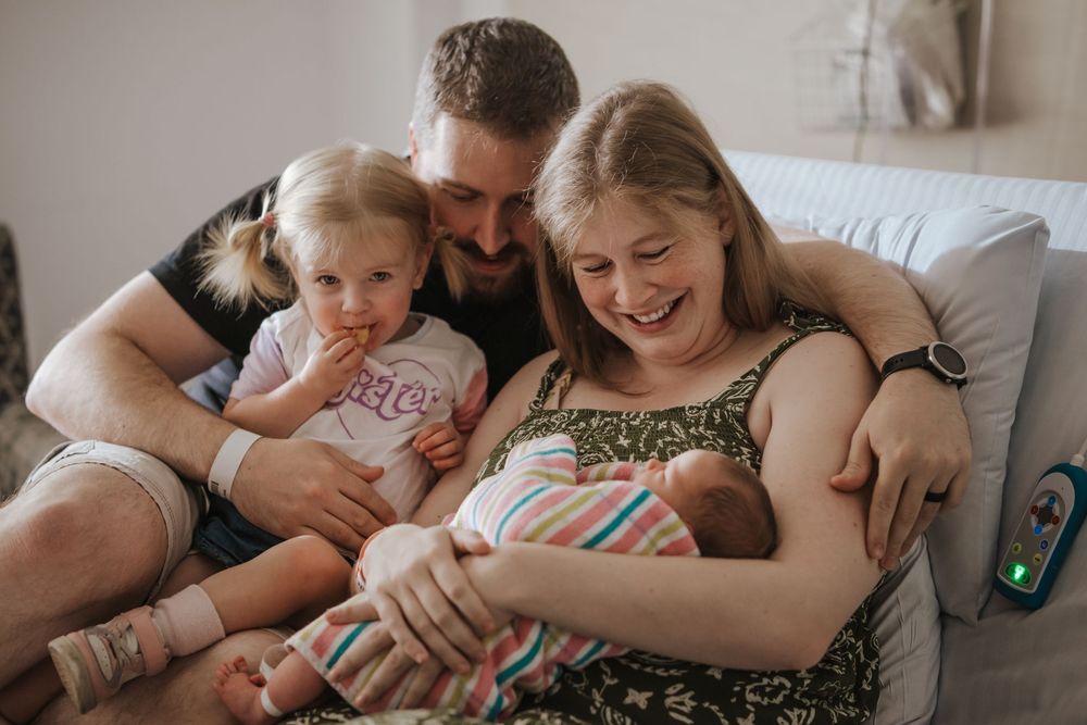 Happy family with dad, toddler girl, and mom holding newborn baby on hospital bed in Norwest after birth.