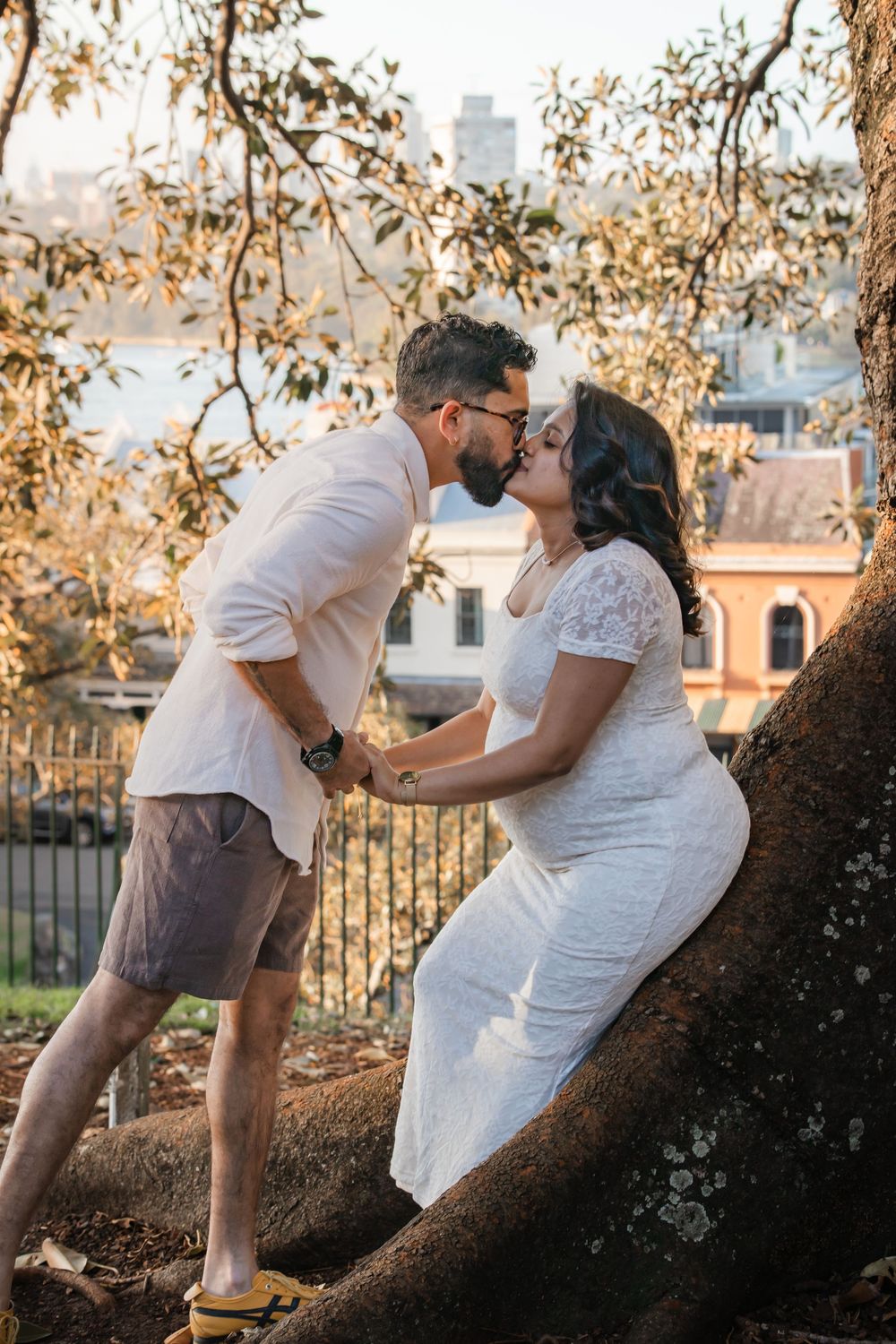 Couple sharing a romantic kiss near a large tree, man in white shirt and shorts, woman in white lace dress, outdoors.