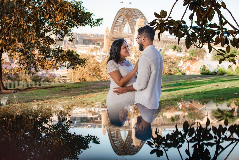 Couple embracing in a park with Sydney Harbour Bridge reflected in a still pond at golden hour.