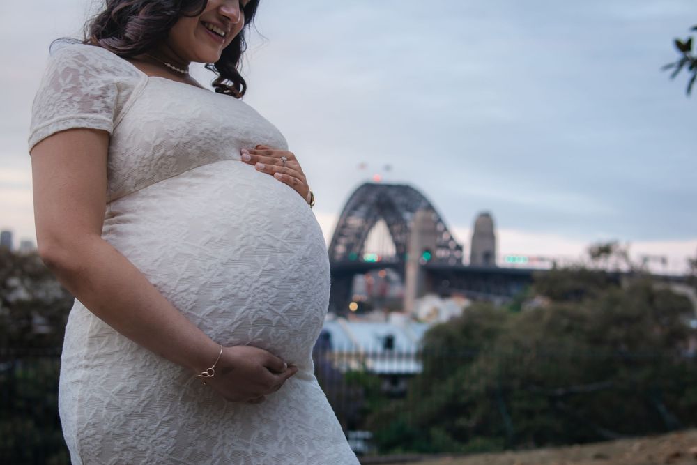 Pregnant woman in white lace dress cradling her baby bump with Sydney Harbour Bridge in the background.