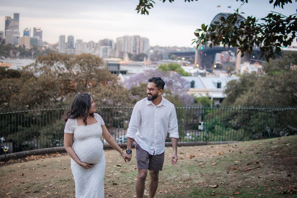 Pregnant couple holding hands walking in park with Sydney city skyline and jacaranda trees in background.