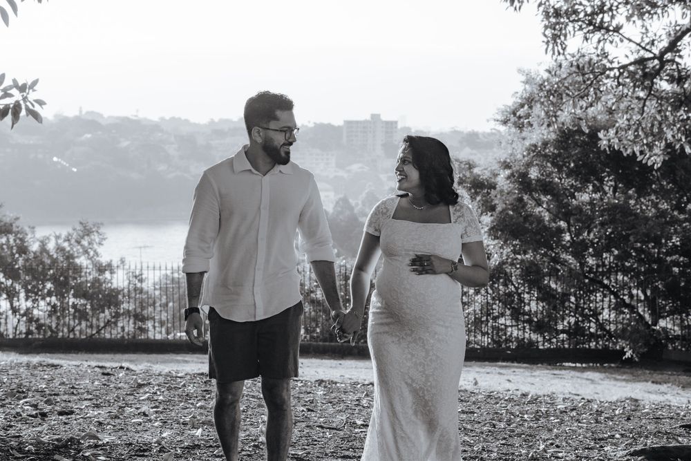 Black and white photo of a couple holding hands outdoors, woman in white maternity dress, man in white shirt, Sydney city backdrop.