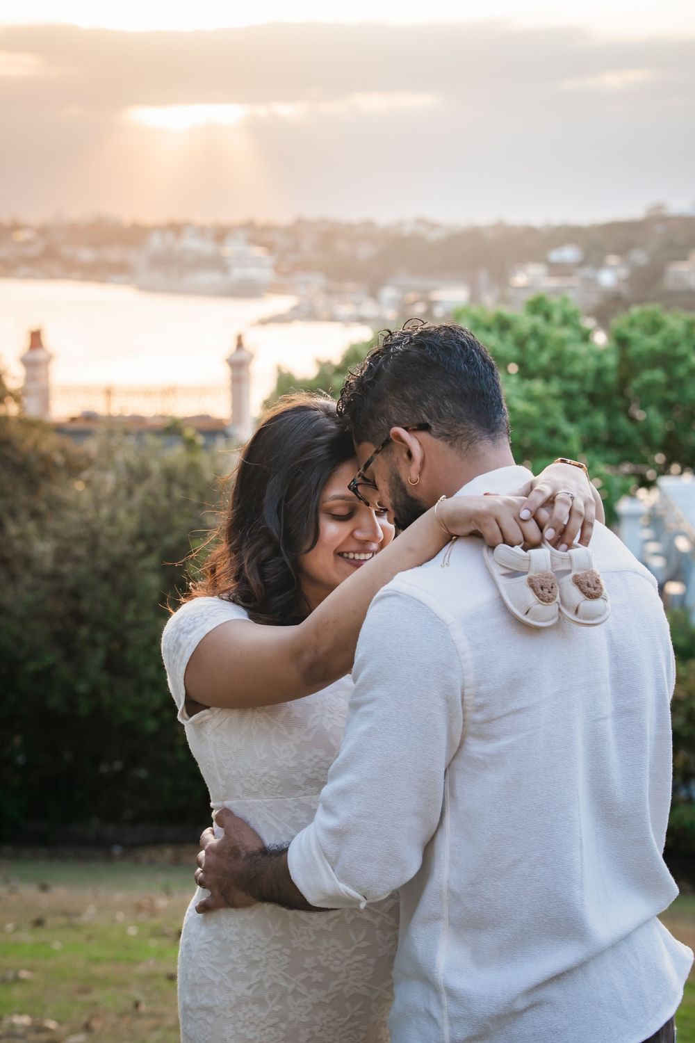 Couple embracing at sunset with Sydney Harbour and lighthouse in background, woman smiling in white lace dress.