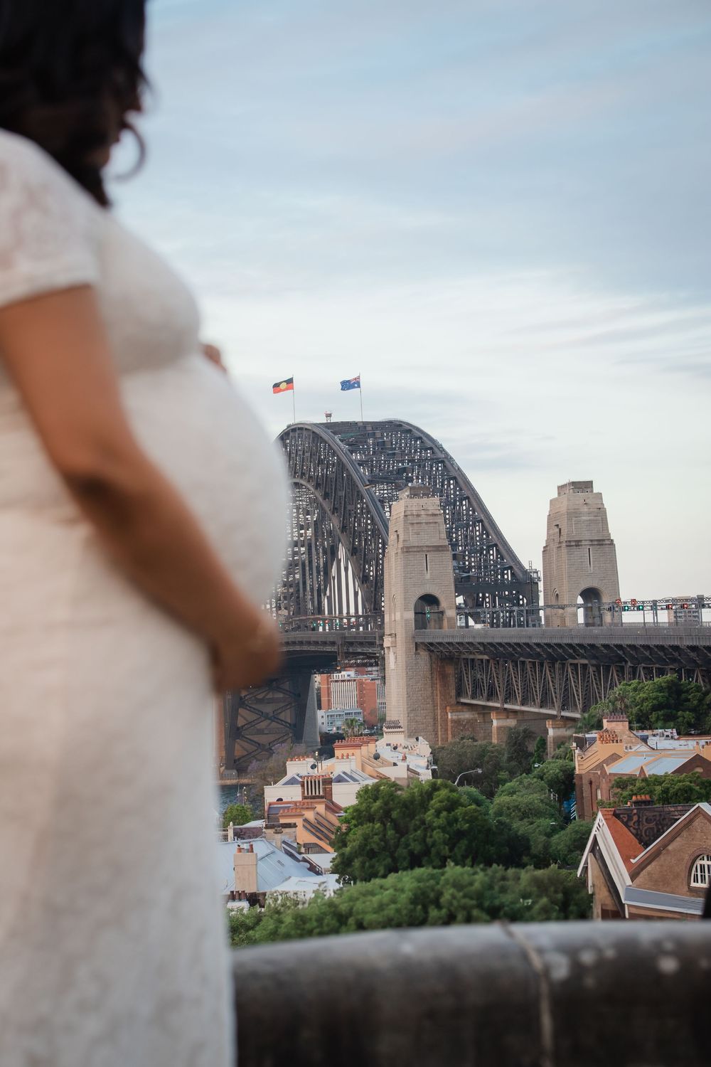Pregnant woman in white lace dress overlooking Sydney Harbour Bridge at dusk.