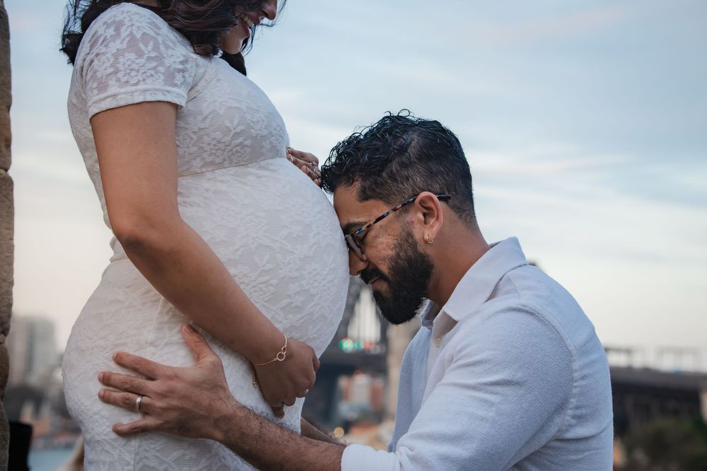 A man kneels to listen to his pregnant partner's belly outdoors during a maternity photoshoot.