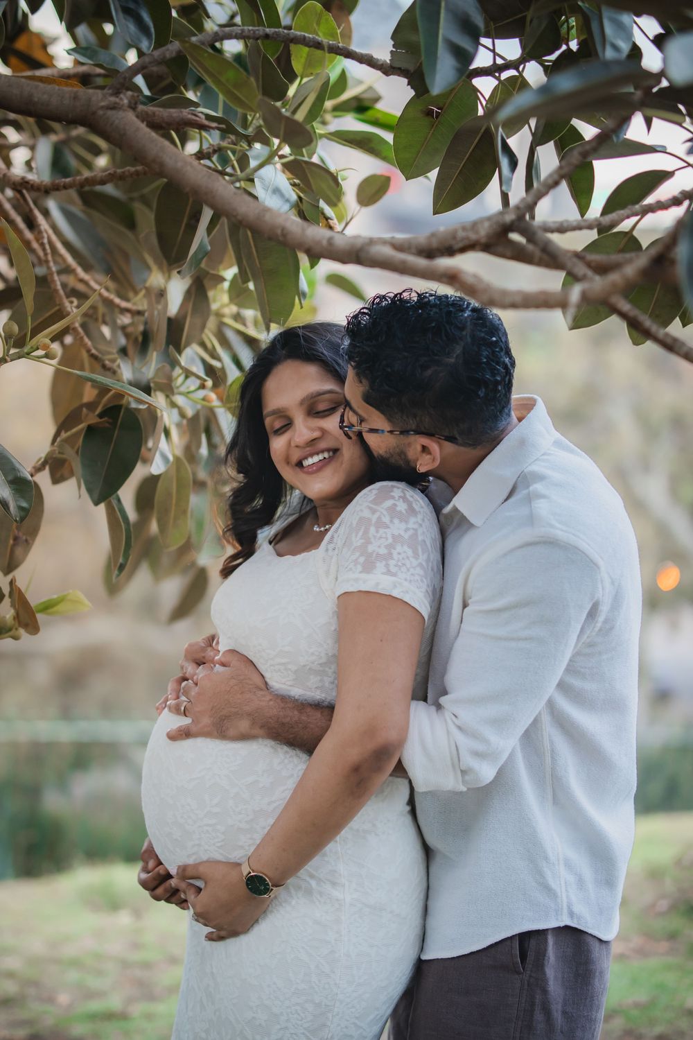 Pregnant couple under tree, man kissing woman's cheek as she smiles, both cradling baby bump in Observatory Hill park.