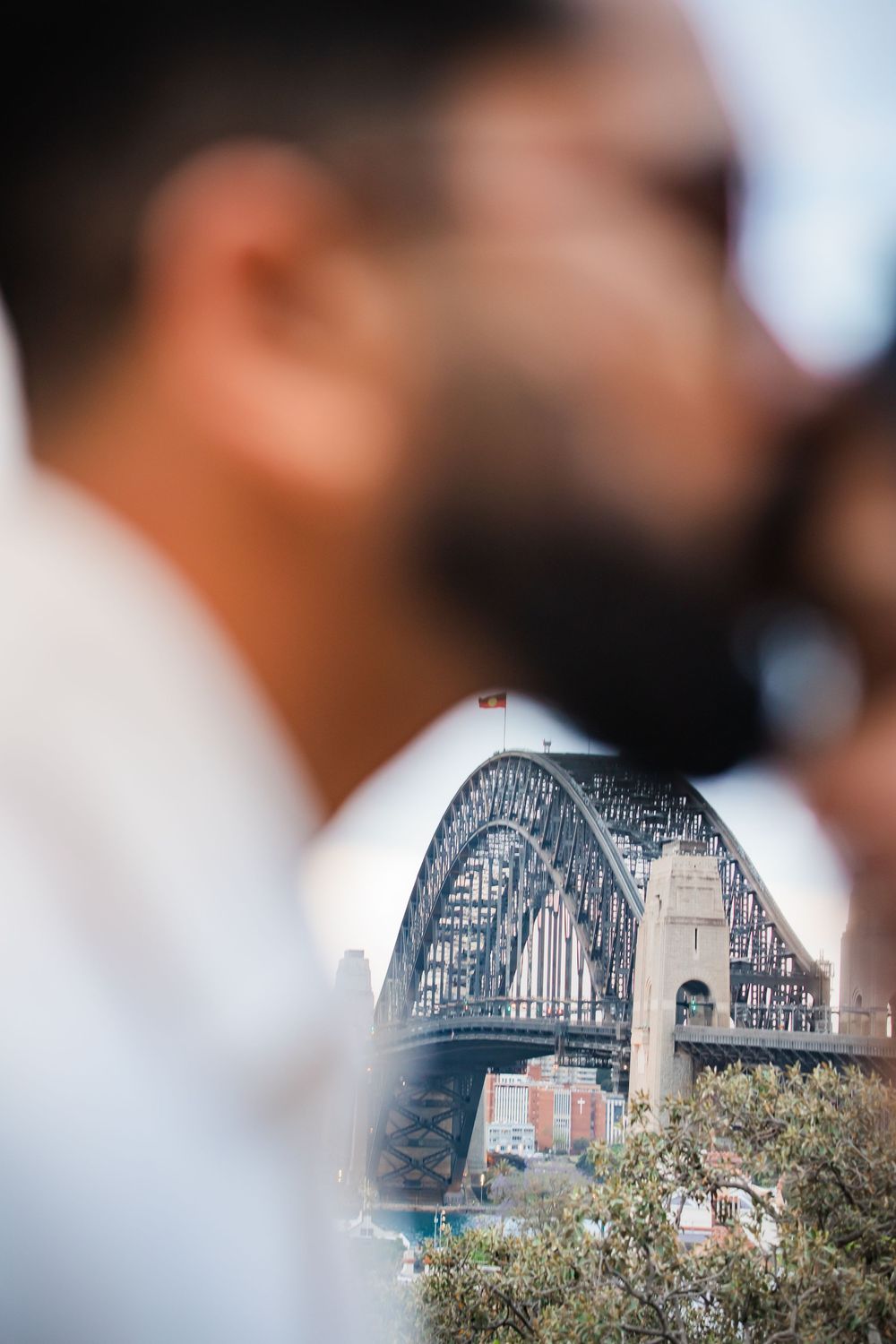 Blurred foreground of a bearded man with Sydney Harbour Bridge in sharp focus between two people.