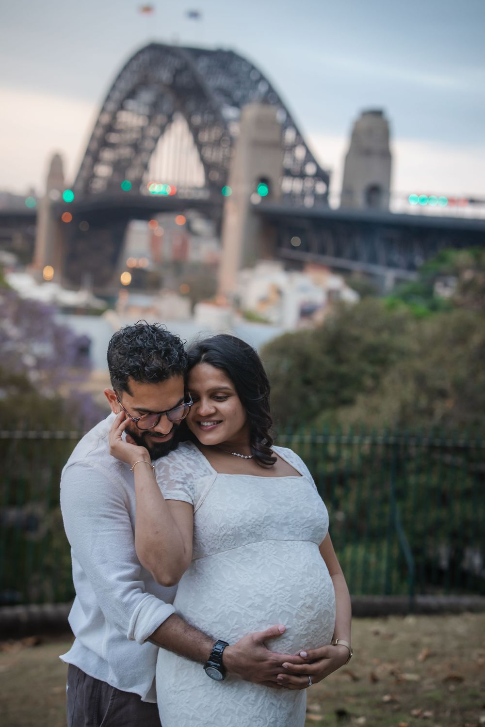 Pregnant couple embracing tenderly in front of Sydney Harbour Bridge at dusk, woman in white lace maternity dress.