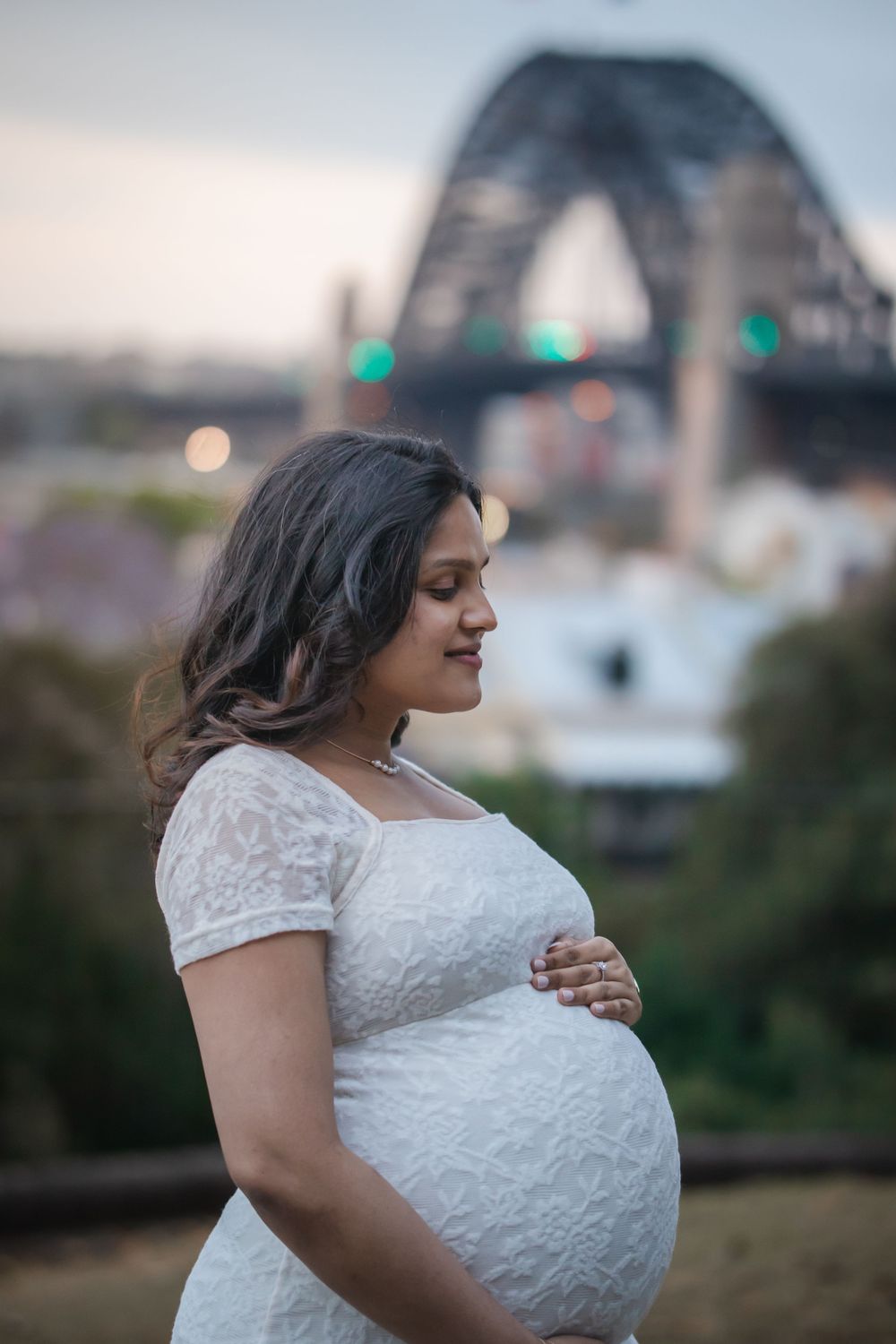 Pregnant woman in white lace dress cradling her bump with Sydney Harbour Bridge in the background at dusk.