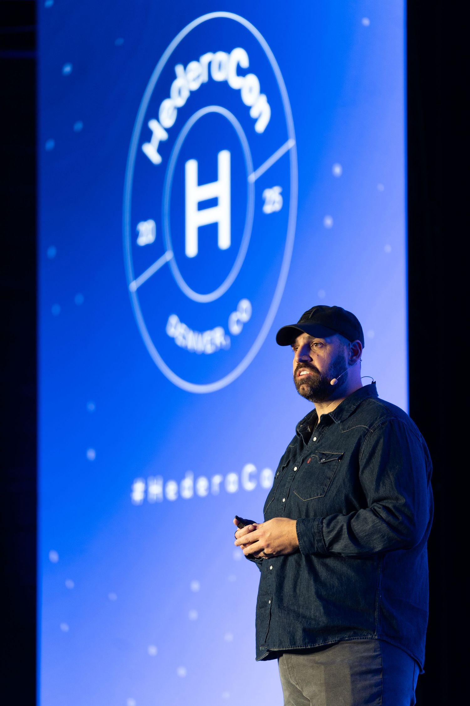 A bearded speaker presents on stage at HederaCon in Denver, CO, with a blue logo projected behind him.