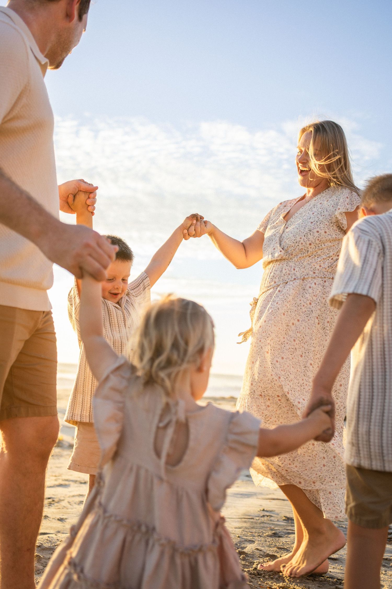 Family holding hands in a circle on a sunny beach at golden hour, with children and parents dancing together.