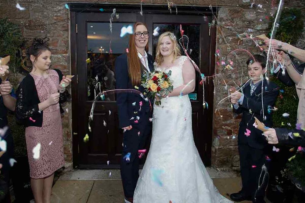 Two brides exit wedding venue as guests celebrate with colorful confetti and wands in joyful ceremony.