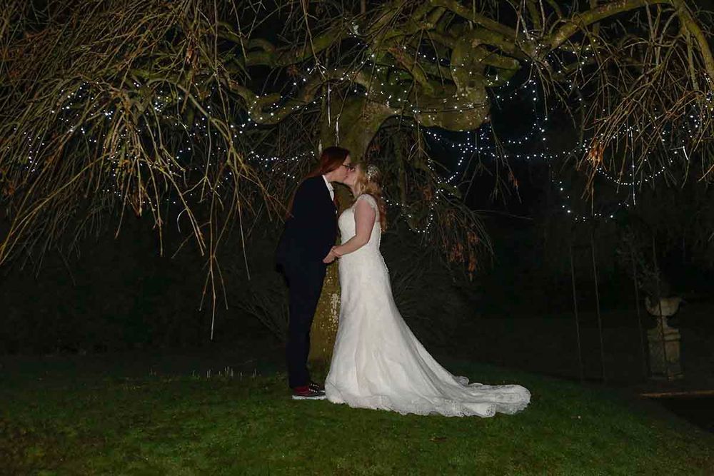 Bride and groom sharing a romantic kiss under a fairy light-adorned weeping willow tree at night.