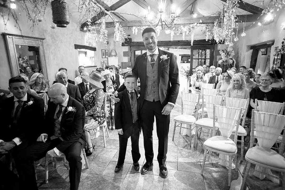 Black and white wedding ceremony photo showing groom and young boy standing at altar in elegant chandelier-lit venue.