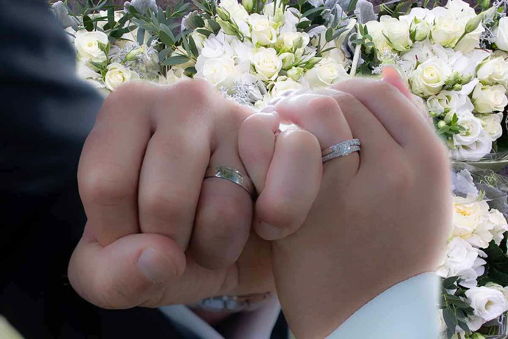 Newlywed couple holding hands with matching wedding rings, with a white floral bouquet in the background.