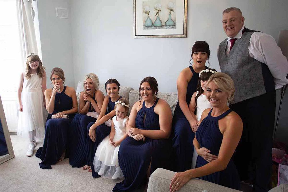 Bridesmaids in navy blue halter dresses pose with a flower girl in white and a man in a vest for a wedding photo.
