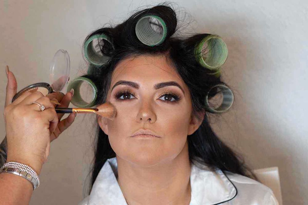 A makeup artist applies foundation with a brush to a woman with dark hair in green velcro rollers wearing a white robe.