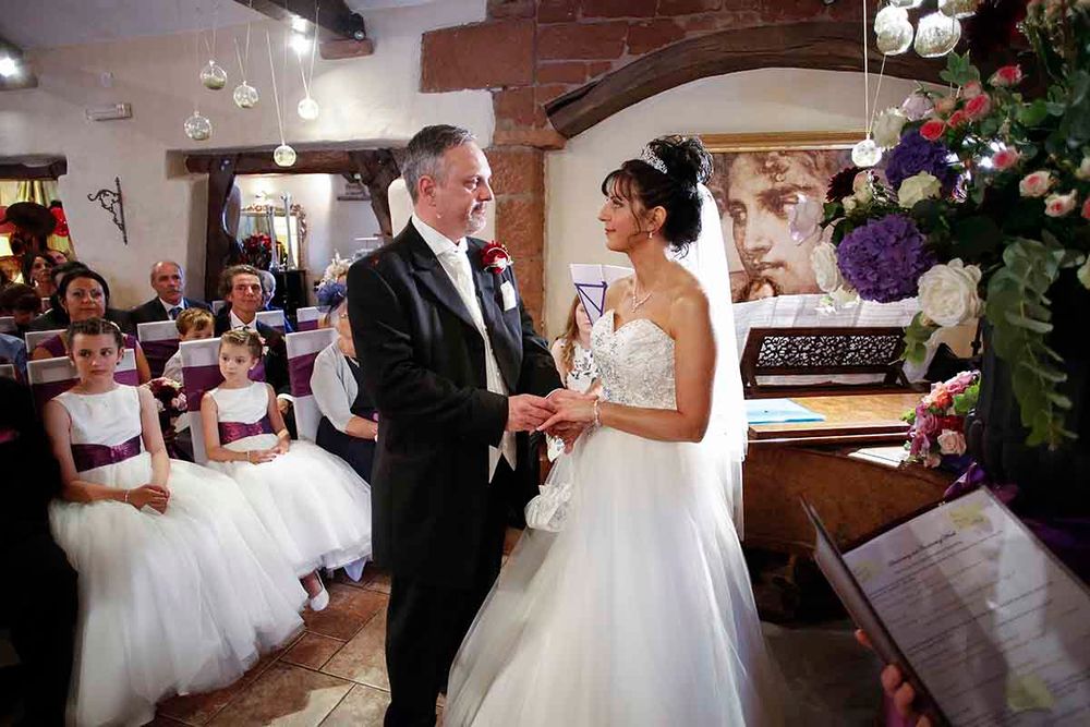Bride and groom exchange vows at elegant indoor wedding ceremony with flower girls seated in background.