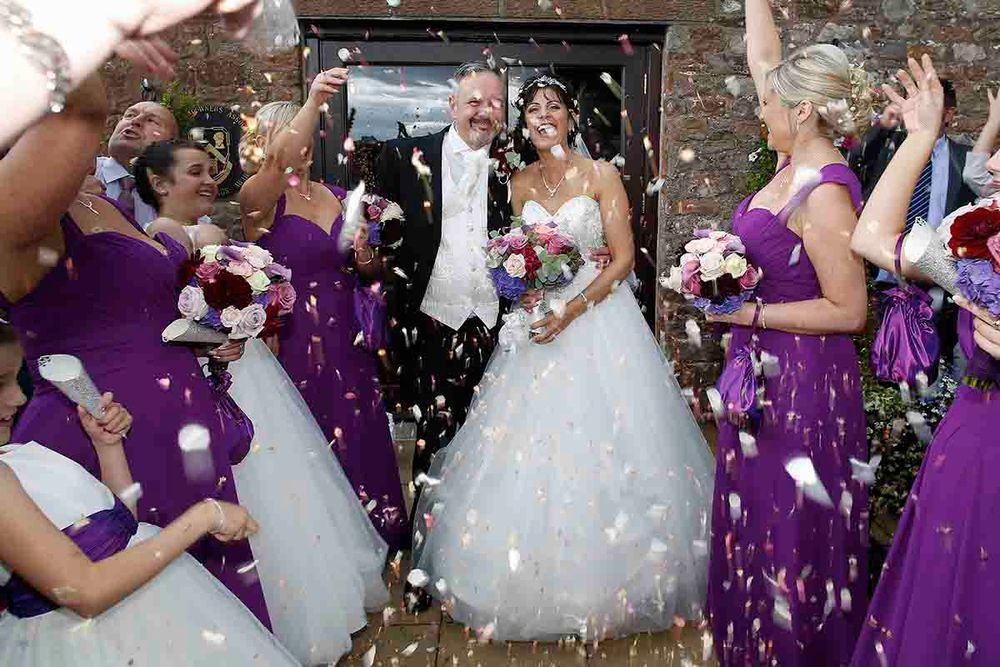 Bride and groom walk through confetti shower surrounded by bridesmaids in purple dresses at outdoor wedding ceremony.