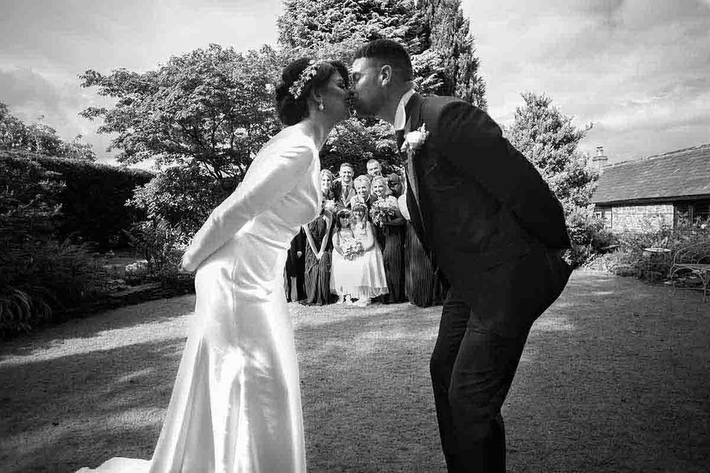 Black and white photo of a bride and groom sharing a kiss outdoors with wedding guests gathered behind them.
