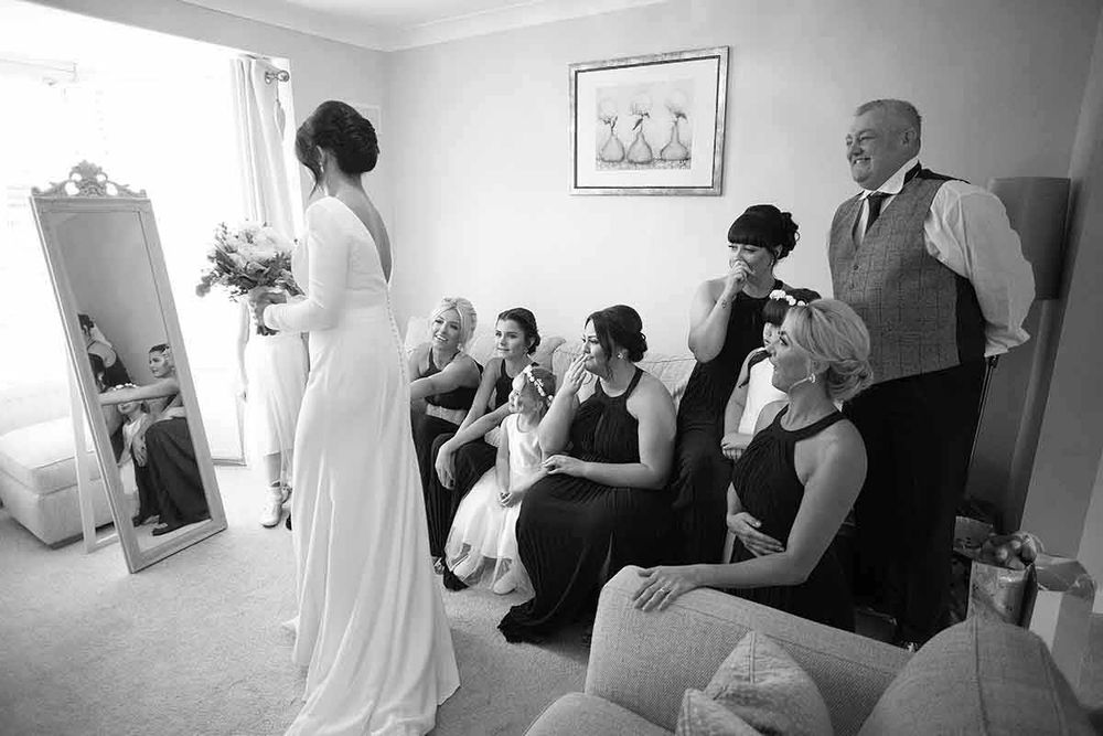 Black and white photo of a bride in a white gown showing her wedding dress to bridesmaids and a groomsman in a bright room.