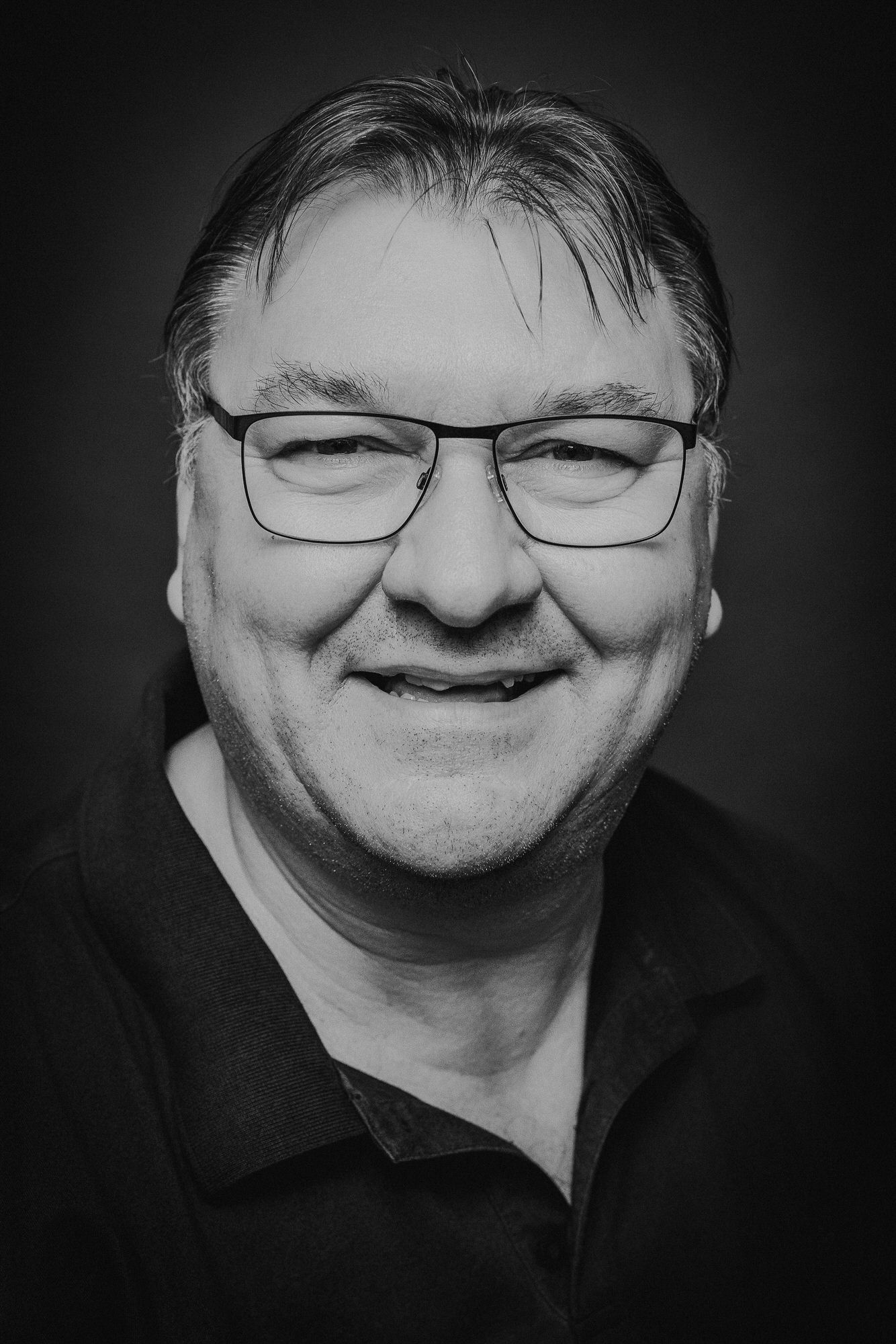 Black and white portrait of a smiling middle-aged man wearing rectangular glasses and a dark polo shirt.