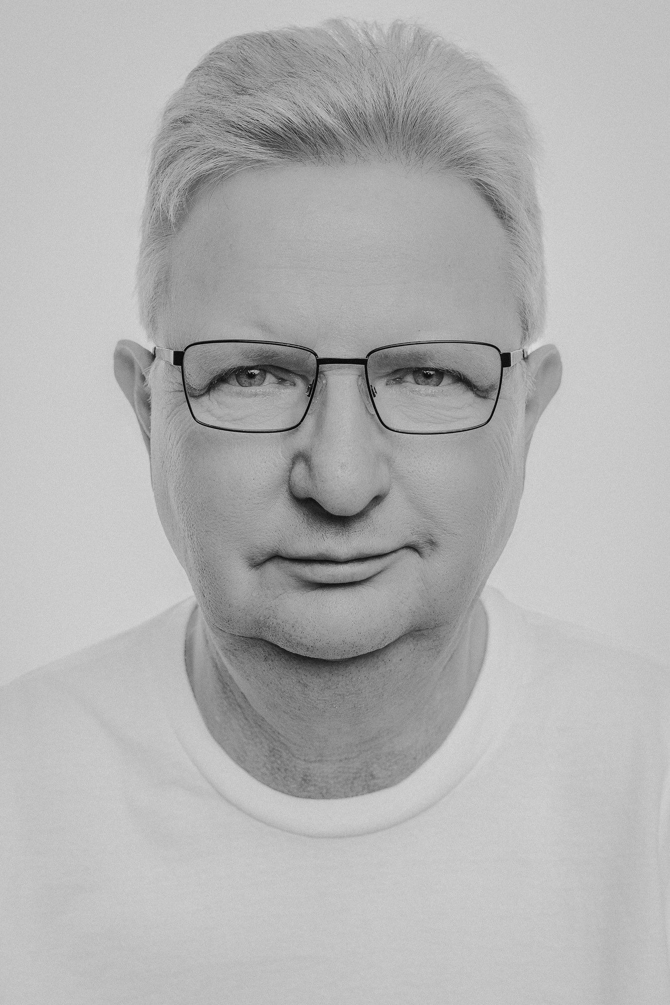 Black and white portrait of a middle-aged man with gray hair wearing rectangular glasses and a white t-shirt.
