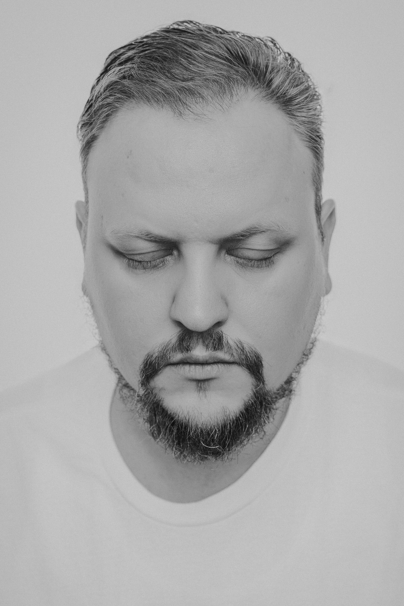 Black and white portrait of a bearded man with closed eyes wearing a white t-shirt against a light background.