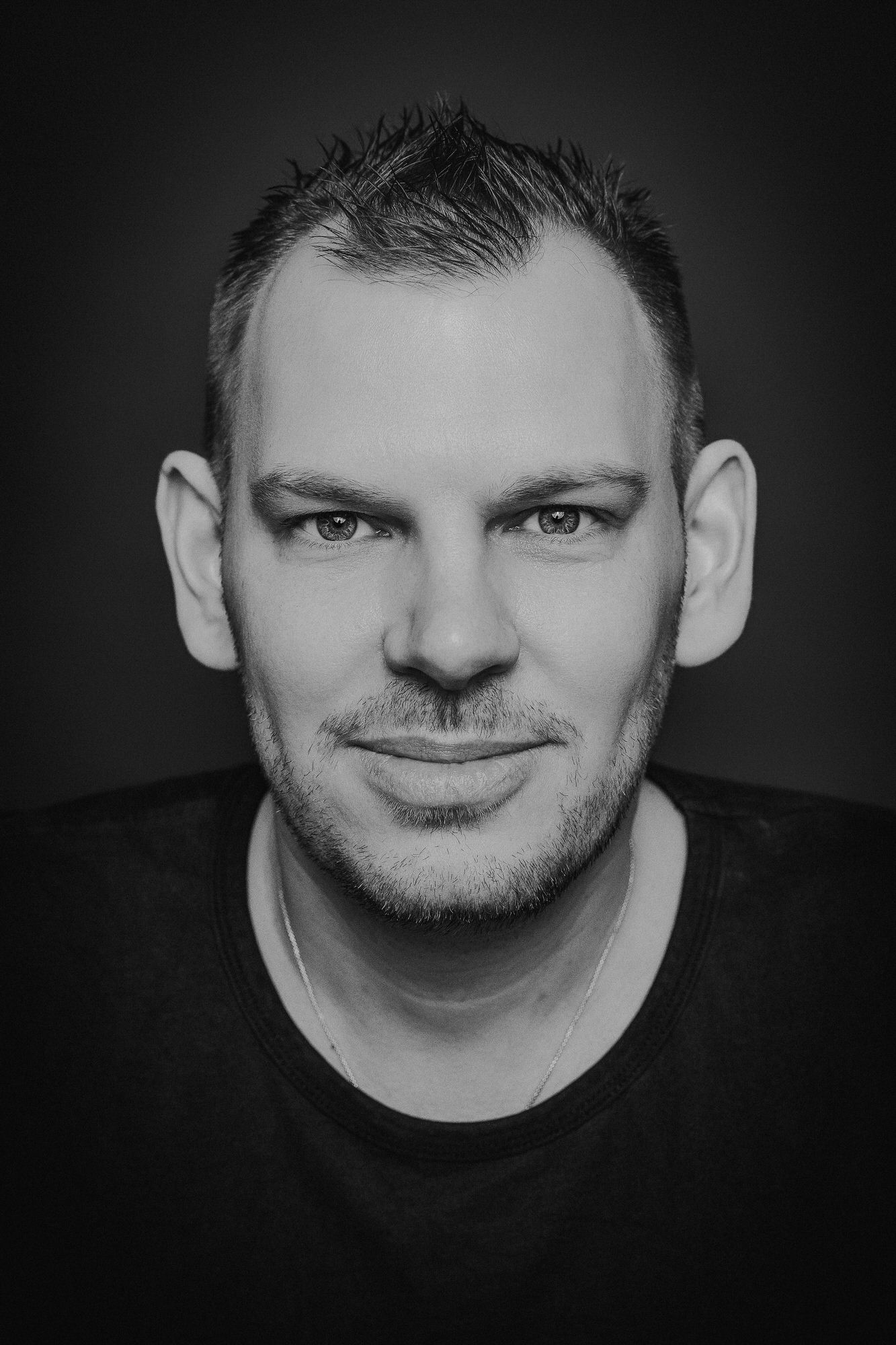 Black and white portrait of a man with short hair and stubble, wearing a dark shirt, against a dark background.