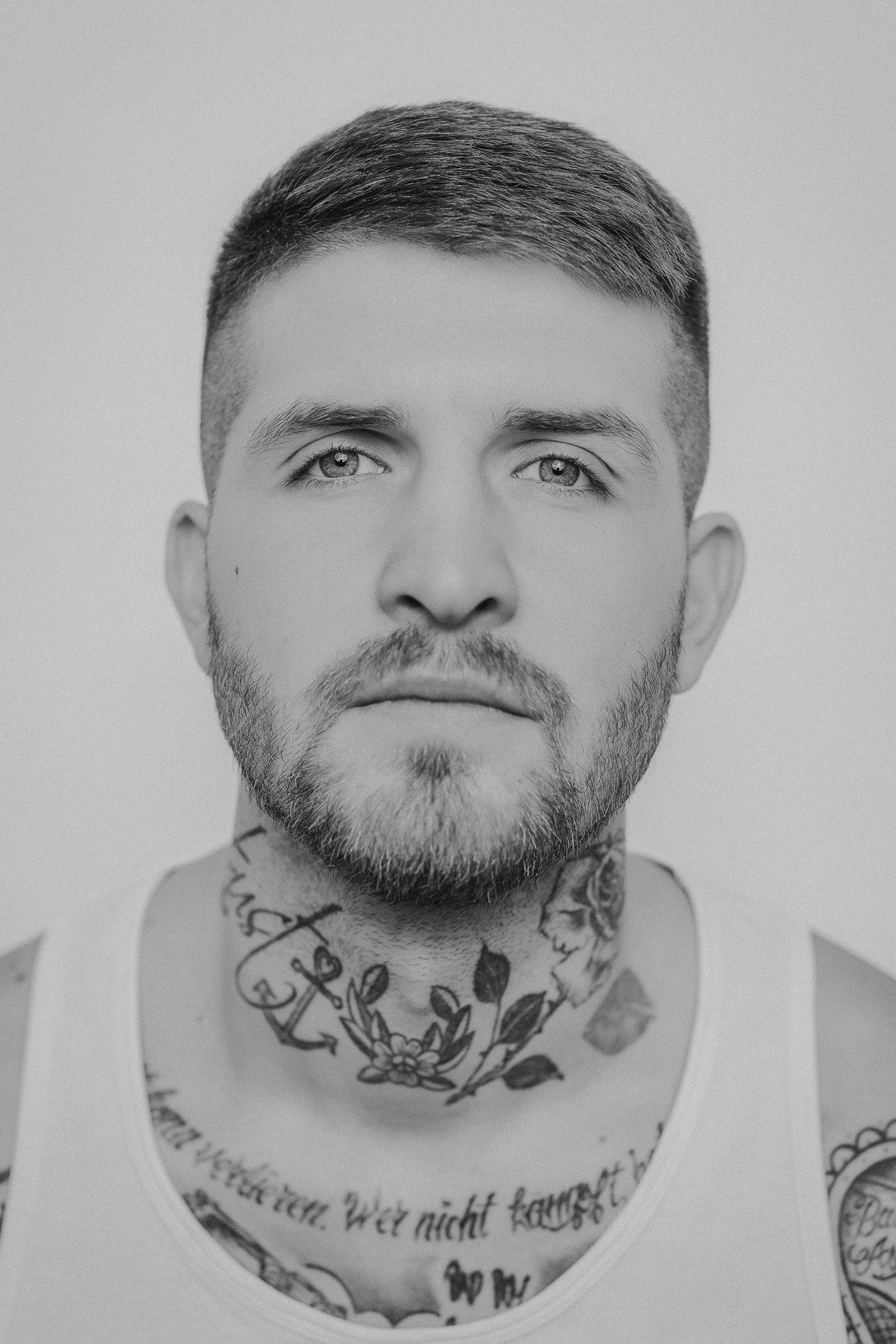 Black and white portrait of tattooed man with beard and neck tattoos wearing a white tank top.