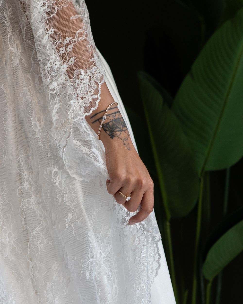Close-up of bride's lace wedding dress sleeve with tattoo and diamond ring visible against tropical foliage.