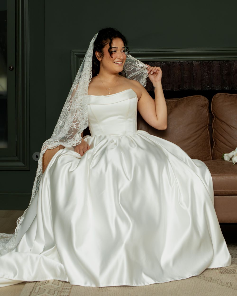 Bride in white satin ball gown with lace veil sitting on brown leather sofa, smiling joyfully.