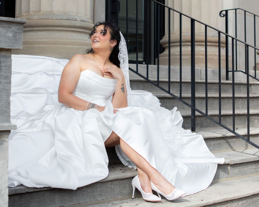 Bride in strapless white wedding gown sitting on steps, showing white heels and lace veil.