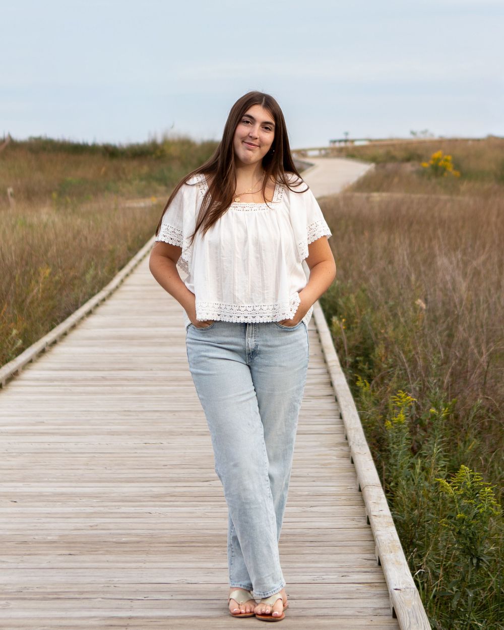 Young woman in white top and light jeans posing on a wooden boardwalk surrounded by coastal grasslands.