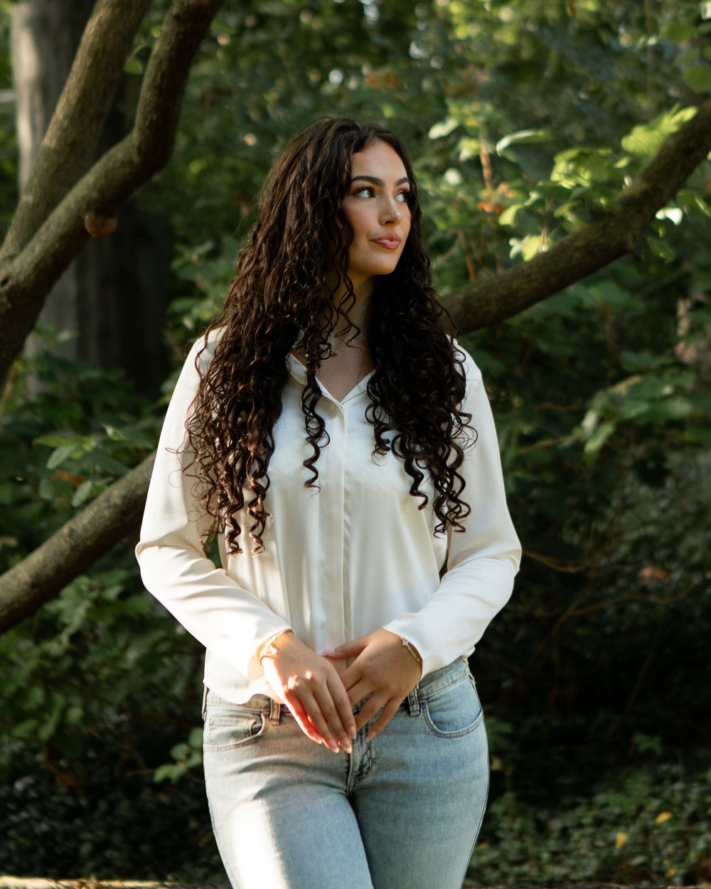 Young woman with long curly dark hair wearing white blouse and jeans standing outdoors among green trees.