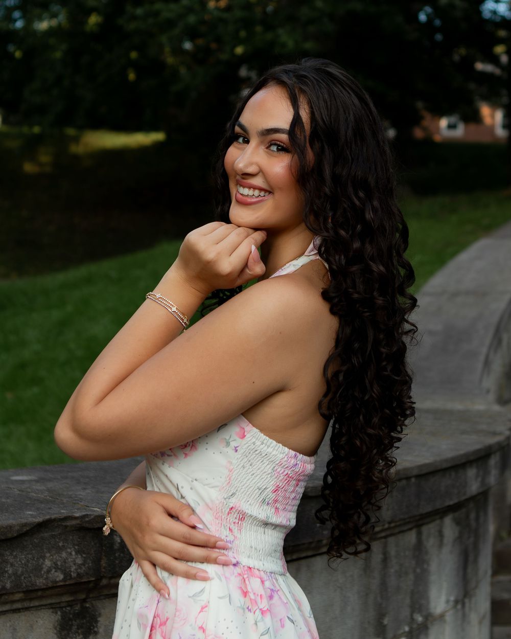 Young woman with long curly dark hair smiles over shoulder in floral dress, posing outdoors near stone railing.
