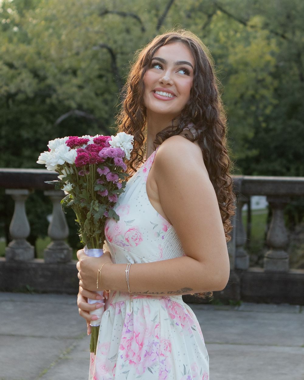 Young woman in floral dress holding pink and white flowers, smiling outdoors near stone balustrade.