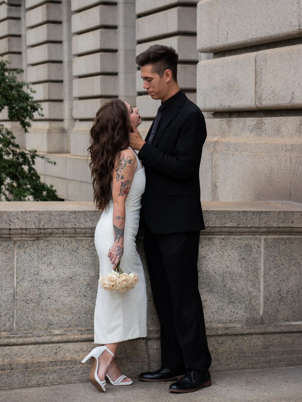 A tattooed bride in a white dress gazes up at her groom in a black suit against a stone wall.
