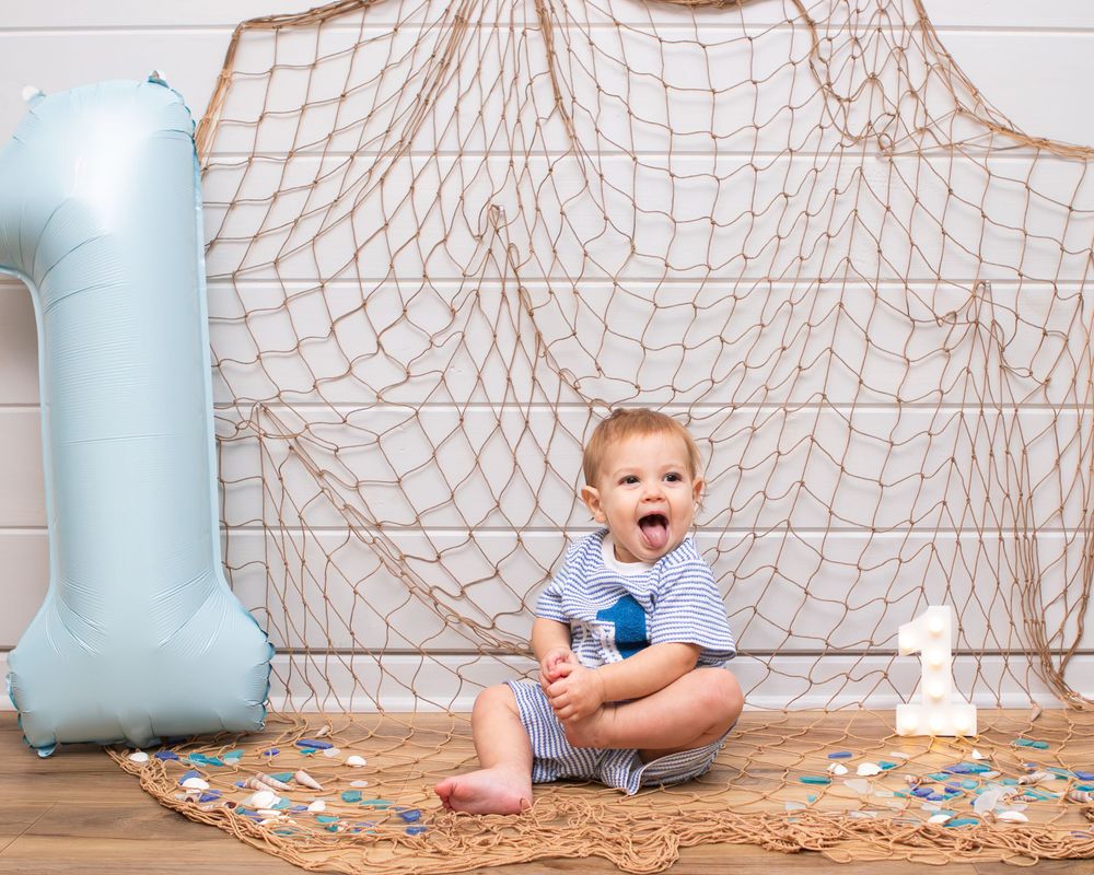 Baby boy celebrates first birthday with blue number one balloon and fishing net backdrop.