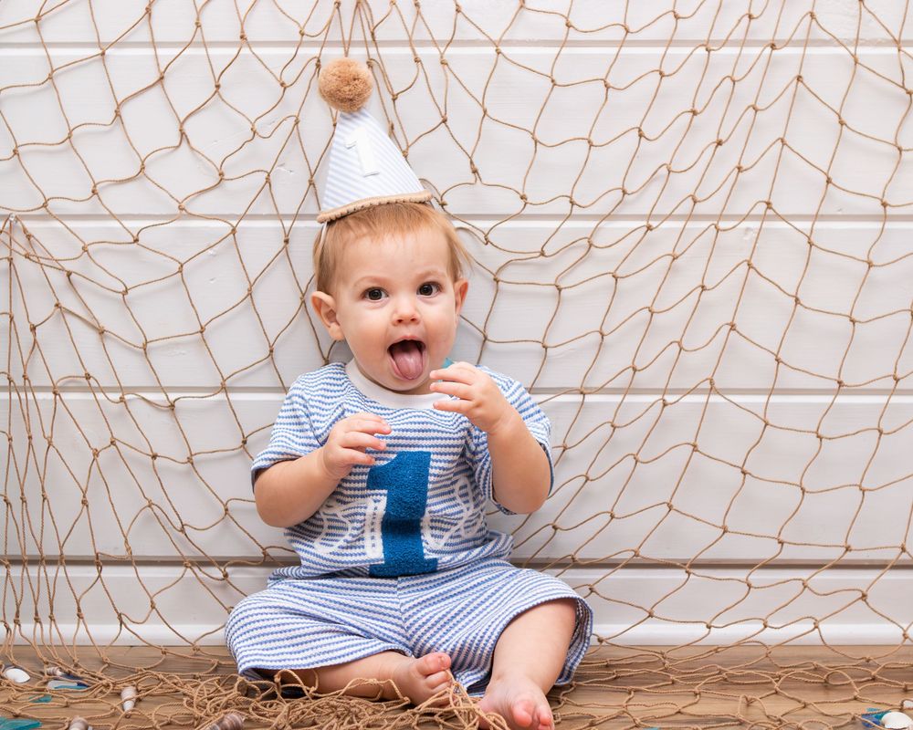 A joyful baby in a striped outfit and party hat sits on the floor holding a number one, celebrating their first birthday.