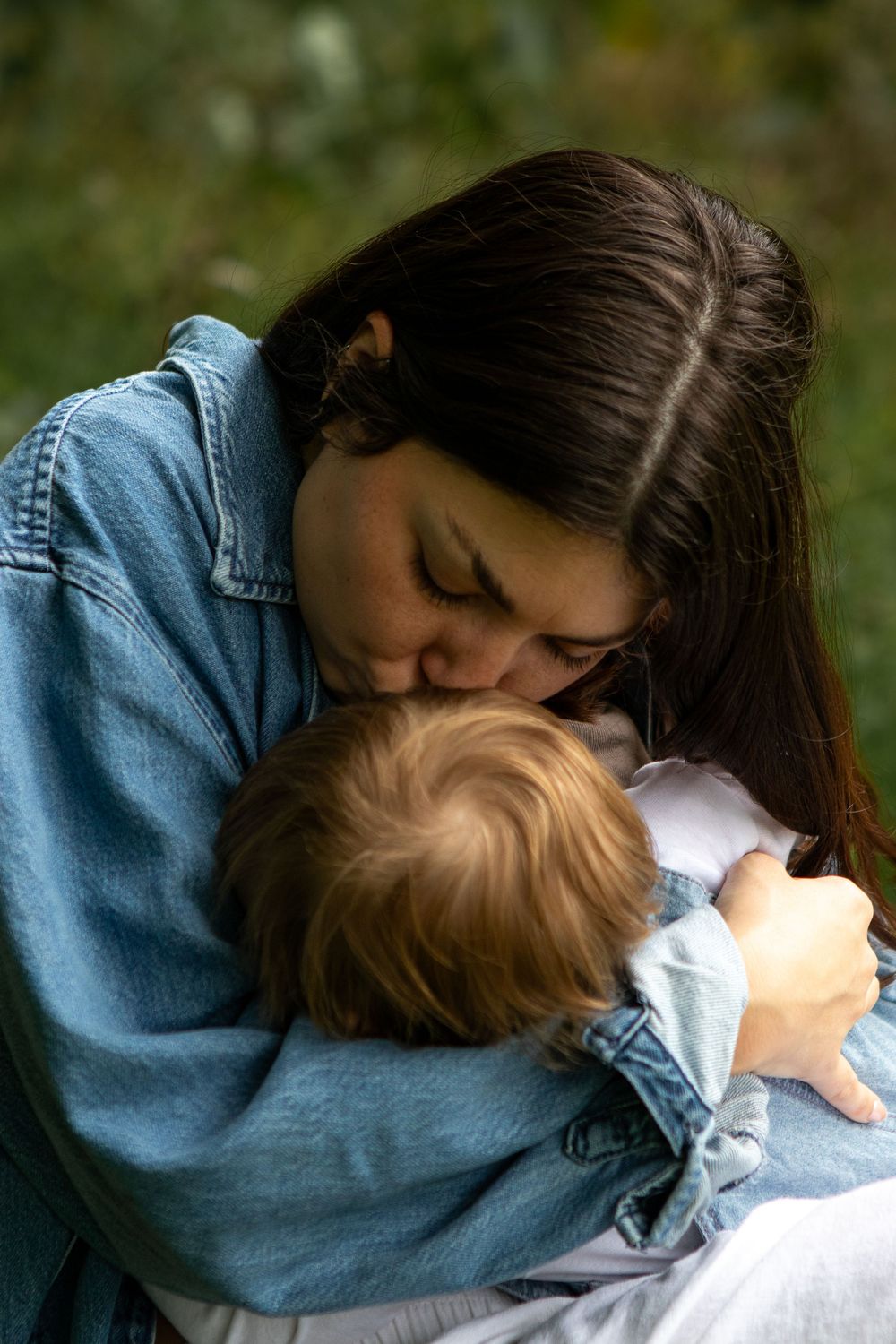 A young mother in a denim jacket tenderly kisses her baby outdoors with a green nature background.