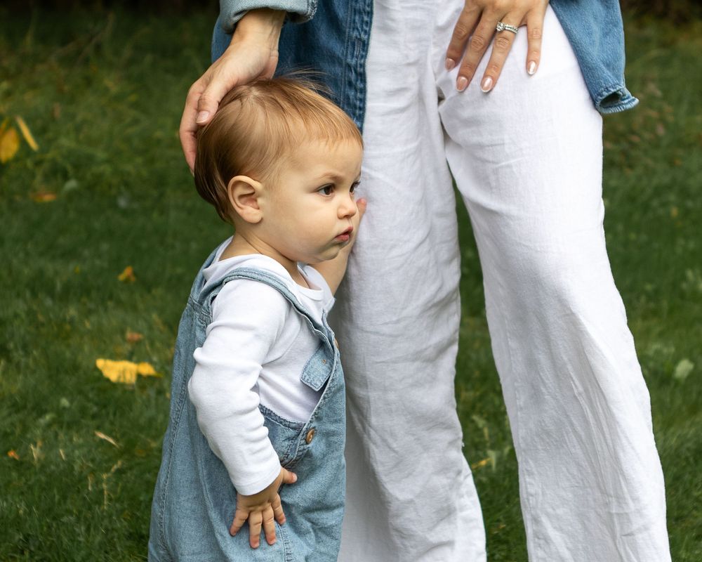 Toddler in denim overalls clinging to mother's leg outdoors on green grass.