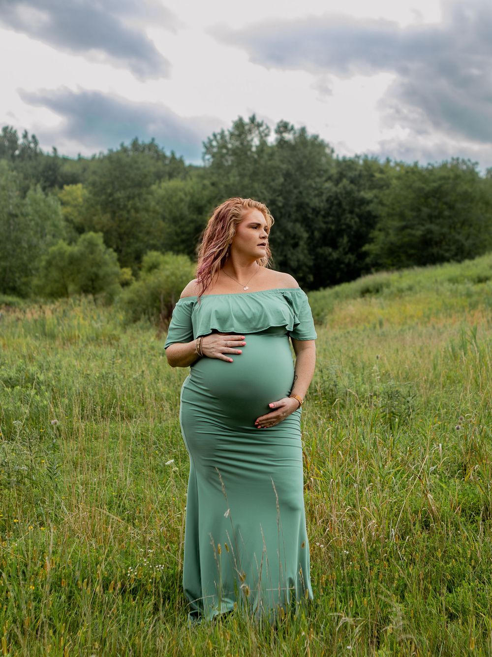 Pregnant woman in sage green off-shoulder gown standing in lush green field under cloudy sky.