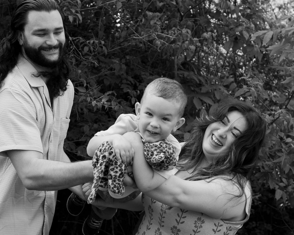 Black and white photo of happy family with curly-haired parents lifting laughing baby outdoors near green foliage.
