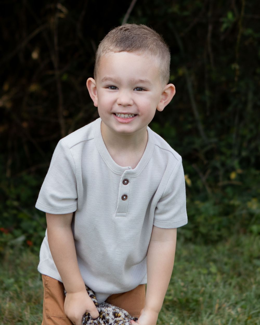 Adorable smiling young boy with short hair wearing gray henley shirt and brown pants posing outdoors in garden.