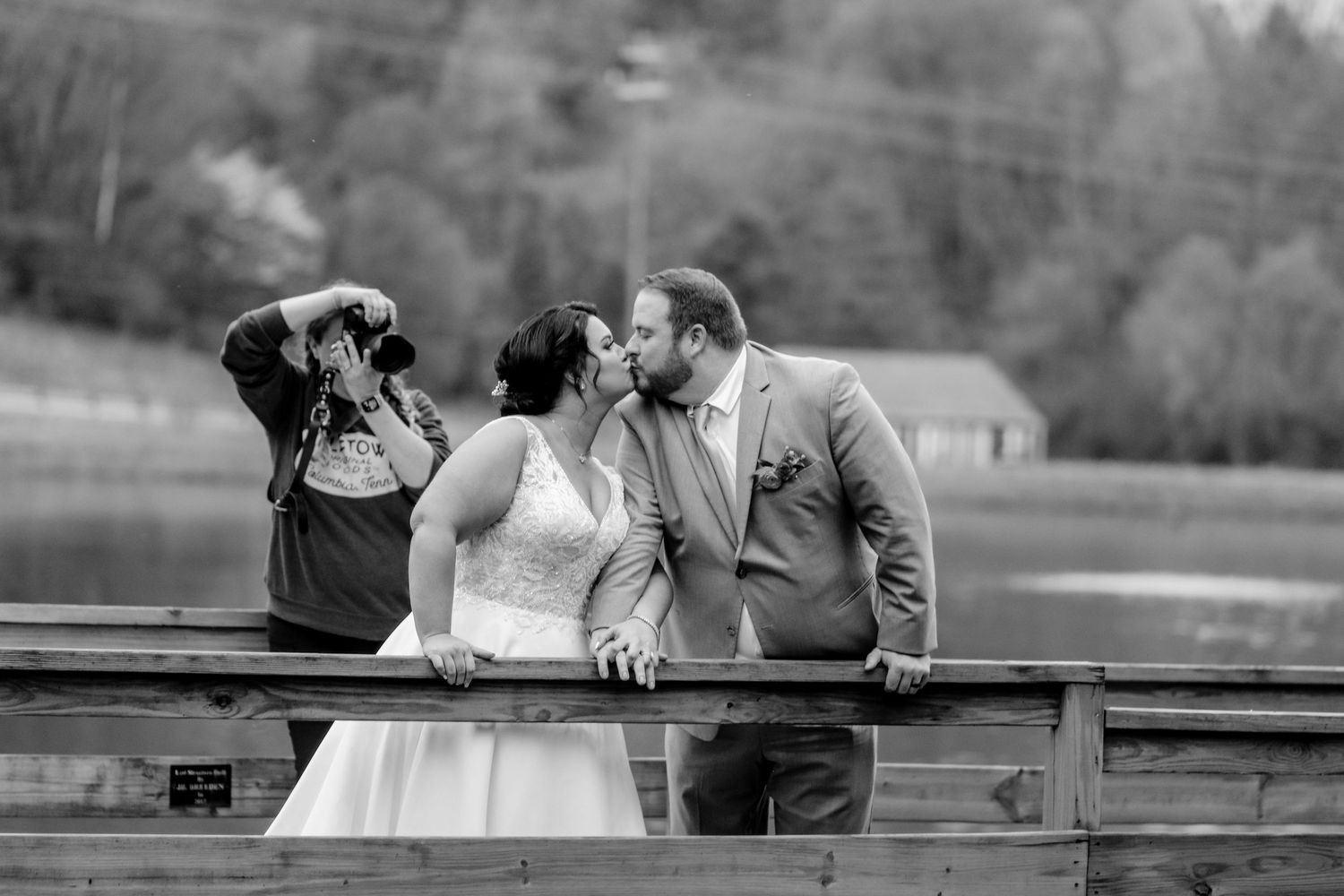 Black and white photo of a bride and groom kissing on a bridge while a photographer captures the moment.