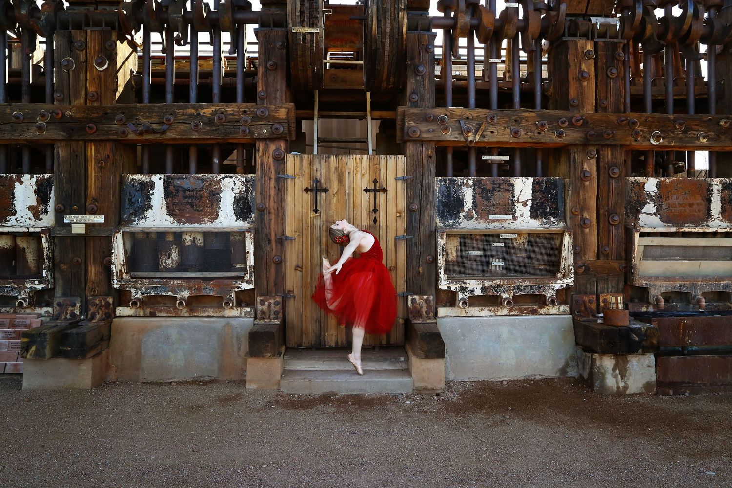 Dancer in a flowing red skirt poses on tiptoe in front of a weathered wooden door at a rustic Western-style building.