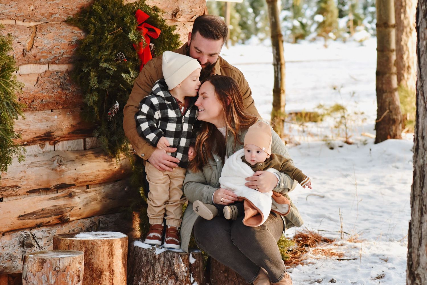Family of four poses outdoors in winter snow near rustic log cabin with holiday wreath decorations.