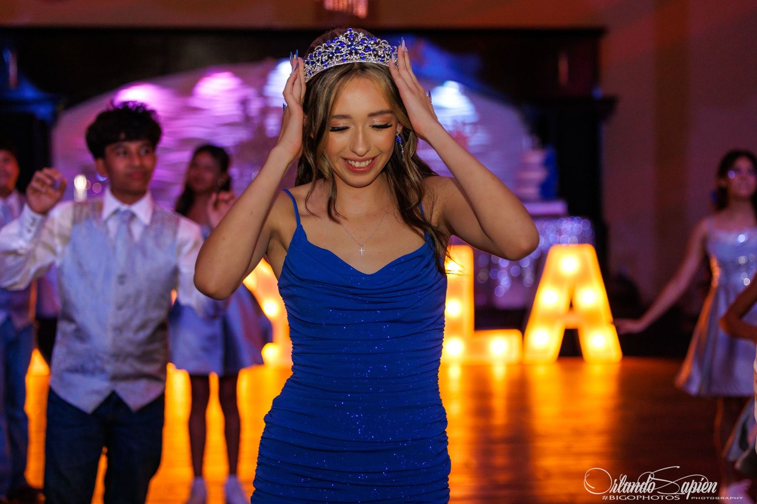 Young woman in blue dress placing tiara on head at quinceañera celebration with dancers and lit letters in background.